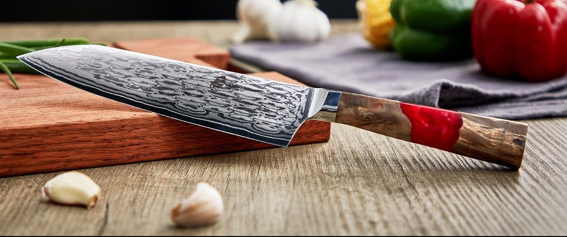 Damascus santoku knife with a wood and red resin handle on a cutting board beside vegetables